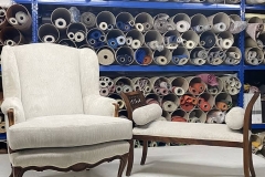 Poltrona bergère e panca coordinate appena restaurate in laboratorio di tappezzeria, con sfondo di rotoli di tessuti pregiati.